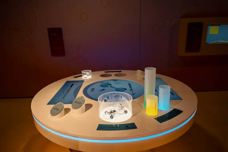 Das Bild zeigt den runden Exponatstisch auf dem unterschiedlich hohe Säulen und drei Bildschirme platziert sind.