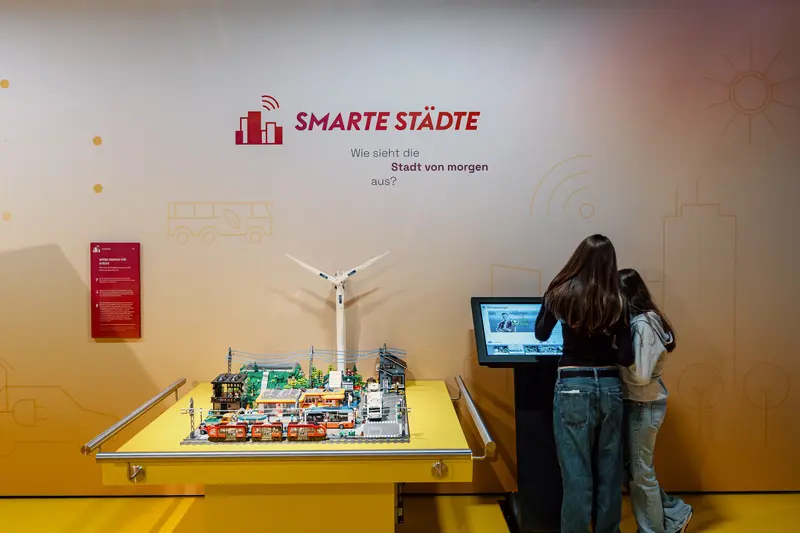 Das Bild zeigt eine Legostadt und zwei Frauen, die sich am Bildschirm daneben Informationen durchlesen.